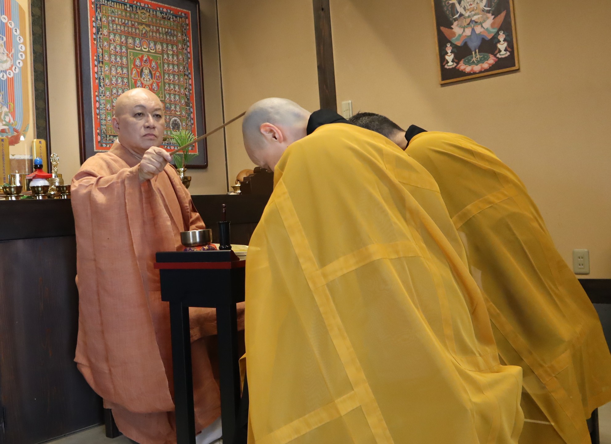寺院の一室で、朱色の衣をまとった阿闍梨（あじゃり）が、洒水器（しゃすいき）と樒（しきみ）の枝葉を使い、黄色い衣の修行僧たちにお加持を行っている厳粛な儀式の様子。壁には曼荼羅（まんだら）などの仏画が掛けられています。